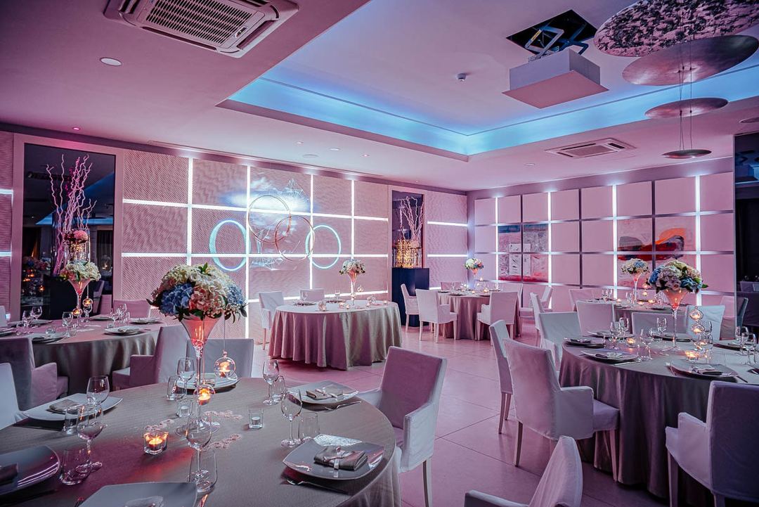 An elegant event space with pink ambient lighting, neon accents, and panoramic windows overlooking the city at dusk.
