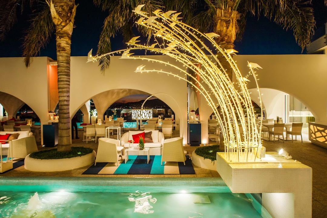 Illuminated poolside lounge area at night featuring cream-colored seating, arched architecture, palm trees, and a decorative lit water feature.