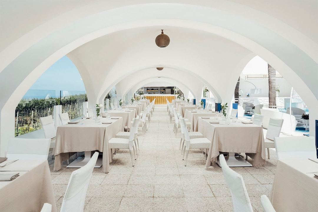 An elegant white arched dining hall with set tables and chairs overlooking a coastal Mediterranean landscape.