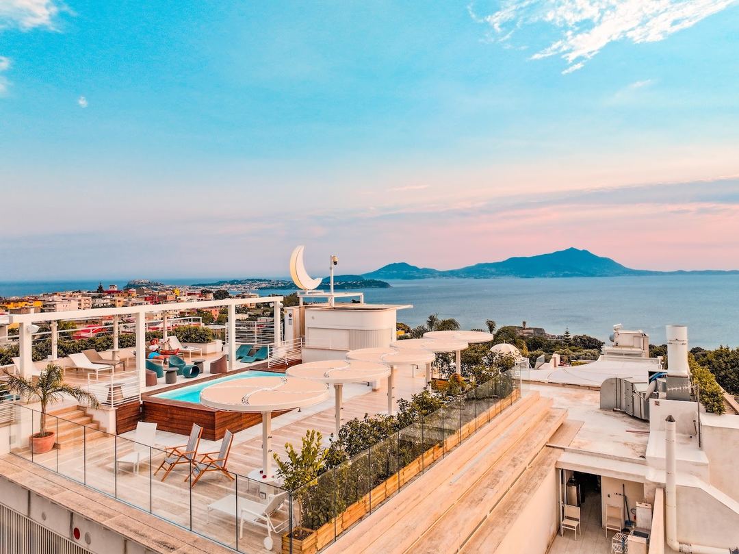 Rooftop terrace with swimming pool, lounging areas, and panoramic views of the coastline and mountains at sunset.