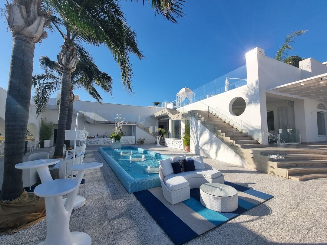 The hotel's poolside area features a bright blue swimming pool surrounded by modern white architecture, palm trees, and comfortable lounge seating under clear blue skies.