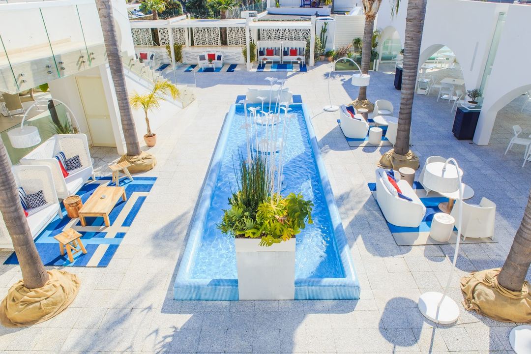 Aerial view of a resort pool area with blue loungers, a narrow lap pool with central planting, palm trees, and white modern architecture surrounding the deck.