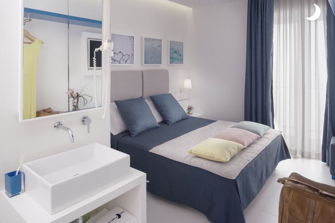 A compact bedroom-bathroom combination featuring a queen bed with blue bedding, integrated ensuite sink, and modern minimalist design.